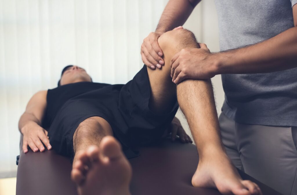 An athlete lying on their stomach with their leg bent upward while a therapist assists with fascial stretch therapy.