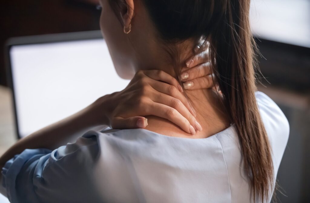 A woman sitting in front of a laptop, holding her neck and shoulder in discomfort.