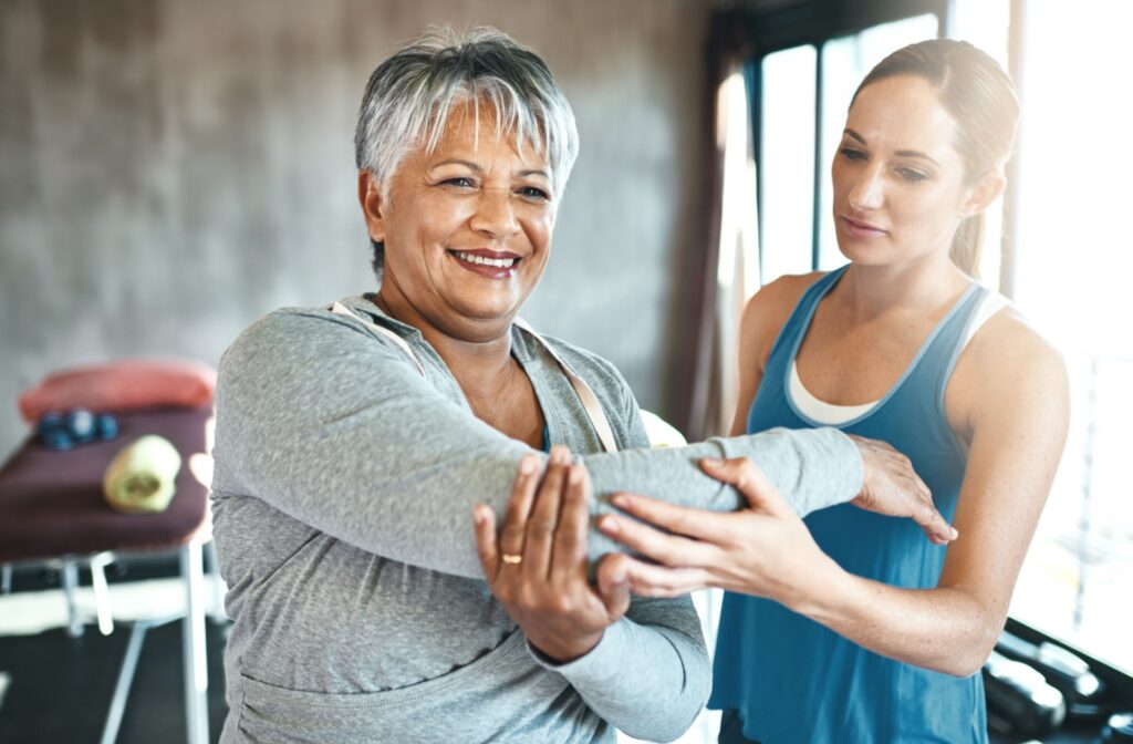 Older patient smiling while doing a light stretching exercise with physiotherapist observing and guiding form.
